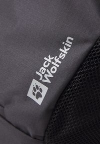 Tessuto grigio con texture verticale, caratterizzato da un logo bianco "Jack Wolfskin" e un'icona di zampa, insieme a una sezione in rete nera.