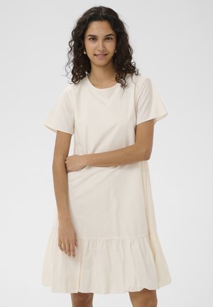 Jeune femme aux cheveux bouclés portant une robe beige clair à manches courtes avec un ourlet à volants, debout, un bras croisé sur son corps.