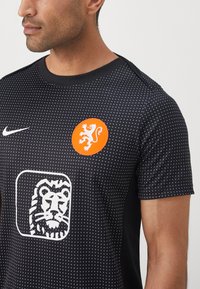 Svart Nike t-skjorte med prikket tekstur, med et oransje løveemblem og en hvit løvegrafikk. Kortermet og med rund hals-design.