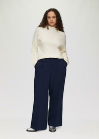 Cremefarbener Strickpullover mit hohem Kragen, kombiniert mit marineblauen, weiten Hosen. Das Model trägt silberne Schuhe. Der Hintergrund ist reinweiß.