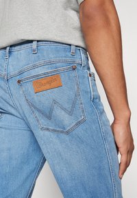 Los jeans de mezclilla azul claro cuentan con una etiqueta de cuero con el logo "Wrangler", costuras en contraste y un patrón de zigzag distintivo en el bolsillo trasero.