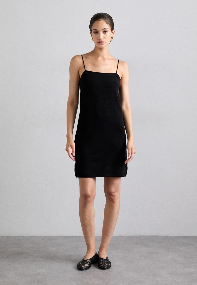 Abito nero a slip con spalline sottili, silhouette dritta, tessuto liscio, lunghezza al ginocchio, abbinato a scarpe nere a slip-on intrecciate.