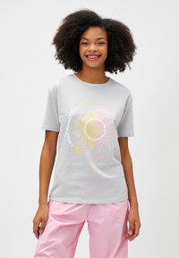 Harmaa puuvillainen t-paita, jossa on keltainen ja vaaleanpunainen auringon sekä yin-yang -grafiikka, ja alla teksti "LÖYDÄ TASAPAINOSI".