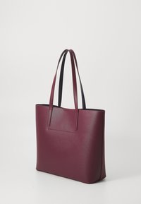 Sac fourre-tout en cuir bordeaux avec texture lisse, forme rectangulaire, doubles poignées et une poche avant discrète. Design minimaliste, sans matériel visible.