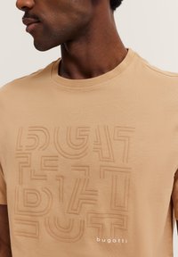 Beige t-shirt with a textured design featuring stylized lettering. Short sleeves, round neckline, and subtle logo at the bottom. Soft fabric.