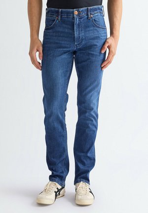 Mężczyzna ubrany w dopasowane niebieskie dżinsy i białe tenisówki na sznurowadła, stojący na tle jednolitego jasnego tła.