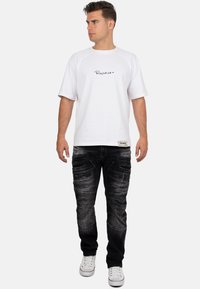 T-shirt blanche à manches courtes avec logo noir, associée à un jean noir déchiré avec poches et baskets blanches. Modèle de profil légèrement tourné.