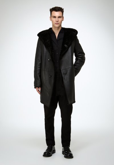 Schwarzer Ledermantel mit großem Kunstpelzkragen, doppelreihigem Design und unstrukturierter Silhouette, kombiniert mit schwarzer Hose und Schuhen.