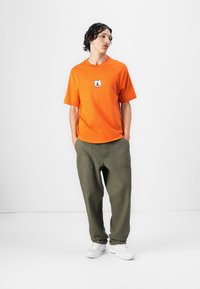 Orange bomulds t-shirt med logo på forsiden, parret med løstsiddende olivengrønne bukser. Modellen bærer hvide sneakers. Ensfarvet baggrund.