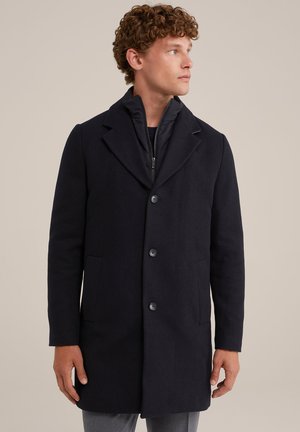 Mann in einem schwarzen, geknöpften langen Mantel über einer Reißverschlussjacke, der nach rechts schaut, vor einem einfachen beigen Hintergrund.