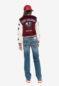 Bordowa i kremowa kurtka varsity z haftem "True Religion" i naszywkami, zestawiona z wyblakłymi niebieskimi dżinsami z detalami na tylnych kieszeniach.