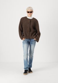 Pull marron, duveteux, boutonné avec des manches longues, associé à un jean bleu clair et des baskets noires. Les accessoires comprennent des lunettes de soleil foncées.