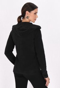 Schwarze Kapuzenjacke mit glatter, texturierter Oberfläche. Lange Ärmel und eine figurbetonte Silhouette. Einfaches Design ohne sichtbare Beschläge oder Verzierungen.