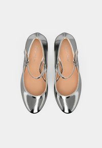 Bailarinas metálicas plateadas con punta redonda, tacón bajo y correa en el tobillo. Textura suave con plantilla beige. Diseño simple.