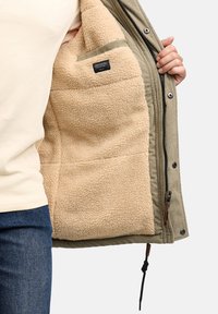 Olivově zelená bunda s měkkou béžovou fleecovou podšívkou a boční kapsou. Má zipové zapínání, manšestrovou texturu a dřevěné knoflíkové detaily.