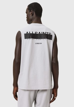 Mann trägt ärmelloses weißes Shirt mit schwarzem Balken und den Worten "ALLSAINTS" und "LONDON" auf der Rückseite, steht vor einem einfarbigen hellen Hintergrund.