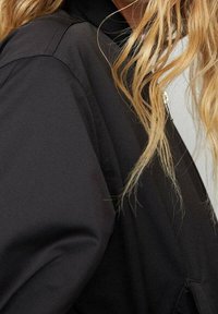 Veste noire avec une finition mate et lisse, dotée d'une fermeture éclair, d'un col montant et d'une conception d'épaule structurée. Cheveux partiellement visibles.