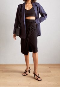 Haut court noir et jupe noire drapée portés sous un blazer bleu marine, associés à des sandales à talons noires. Tissu texturé avec un détail noué sur la jupe.