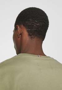 Vista posteriore di una persona con capelli corti e ricci che indossa una camicia verde oliva con un'etichetta a strisce rossa, bianca e blu sul colletto.