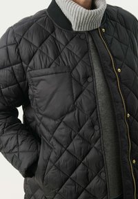 Dekoratiivne must bomber-jope, millel on soonikkoes krae, rinnaehe ja kuldsed nupukinnitusmed. Kihtidena halli kampsuni peal.