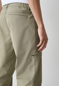 Kaki kravas bikses ar gludu faktūru, elastīgu vidukļa joslu un sānu kabatas detaļām. Tās ir aprīkotas ar aizmugurējo kabatu un piegulošu siluetu.