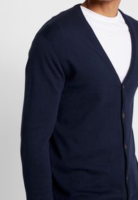 Námořnický modrý cardigan s V-výstřihem, se zapínáním na knoflíky a strukturovanými žebrovanými manžetami. Vyrobeno z měkké a lehké látky.