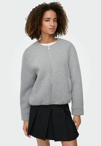 Sweat-shirt gris zippé avec un col rond, des poignets côtelés et des poches latérales, associé à une jupe plissée noire.