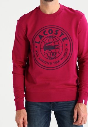 Homme portant un sweat-shirt rouge Lacoste avec un logo bleu marine et un texte indiquant « Lacoste Live All Around The World » sur la poitrine.