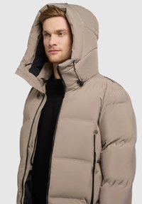 Beige puffer-jack met een hoge kraag en grote capuchon, voorzien van zwarte ritsen en een gladde, glanzende textuur. Zijkanten zichtbaar.