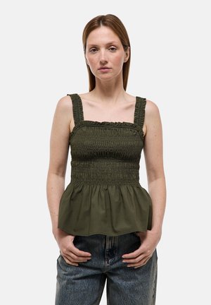 Jeune femme debout, les mains dans les poches, portant un top sans manches froncé vert foncé et un jean bleu sur un fond uni.