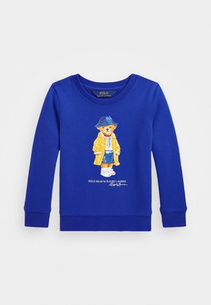 Niebieska bluza z długim rękawem z niedźwiedziem w żółtej kurtce, niebieskiej czapce i tenisówkach, poniżej napis Polo Bear by Ralph Lauren.