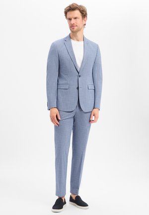 Mann trägt hellblauen Anzug mit Zweiknopf-Blazer und passenden Hosen, weißem T-Shirt und schwarzen Slipper-Schuhen, steht auf weißem Hintergrund.