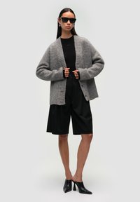 Szary wełniany kardigan o ryfrowanej fakturze i z przodem zapinanym na guziki, noszony z czarnymi szortami o wysokim stanie oraz czarnymi szpilkami, uzupełniony okularami przeciwsłonecznymi.