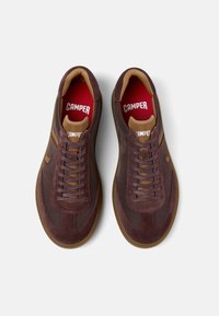 Sneakers din suede maro și plasă, cu talpă din cauciuc, având accente maronii și branțuri roșii, legate cu șireturi prin ochiuri întărite.