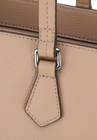 Primo piano di una borsa in pelle beige che mostra una superficie testurizzata, un attacco per manico rettangolare e dettagli di cucitura leggeri. Accenti in metallo argentato inclusi.