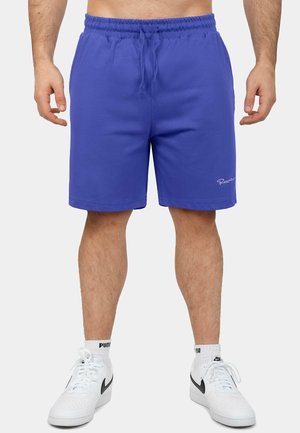 Mann trägt blaue Shorts mit Kordelzug bis zur Mitte des Oberschenkels und weiße Nike-Sneaker mit schwarzem Swoosh sowie weiße Puma-Socken, steht mit entspannten Armen.