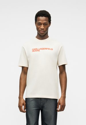 Jonge man die een wit T-shirt met korte mouwen draagt met oranje tekst 'KARL LAGERFELD JEANS', gecombineerd met donkerblauwe spijkerbroek, staand tegen een witte achtergrond.