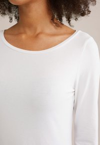 Nahaufnahme einer Person mit lockigem Haar, die ein schlichtes weißes Langarmshirt mit einem weiten runden Halsausschnitt trägt.