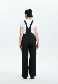 Frau mit schwarzen Haaren im Pferdeschwanz, die schwarze weite Latzhose und ein weißes kurzärmliges Hemd trägt, steht mit dem Rücken zu einem weißen Hintergrund.