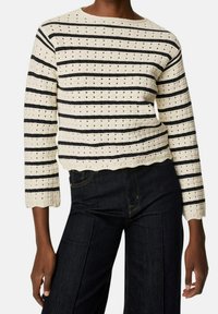 Kremowy sweter z dzianiny w granatowe paski, o strukturze wzoru, z dekoltem w łódkę i rękawami do łokcia, z falowanym wykończeniem u dołu. Noszony z ciemnymi dżinsami.