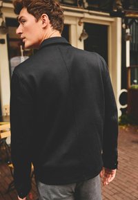 Black, textured jacket with a collar, features a straight cut and buttoned cuffs, shown from the back against a café backdrop.