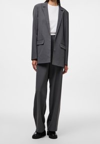 Blazer gris sur mesure avec deux poches avant, assorti à un pantalon à jambes larges pareil. L'ensemble présente un tissu lisse et texturé.