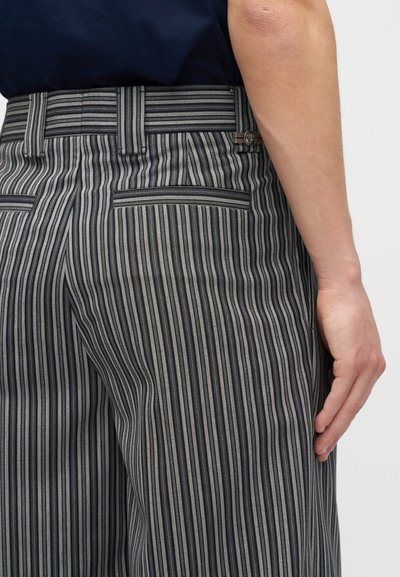 Κοντινή λήψη ενός ατόμου που φοράει μαύρο και γκρι παντελόνι με κάθετες ρίγες, με το δεξί του χέρι χαλαρά στο πλευρό.