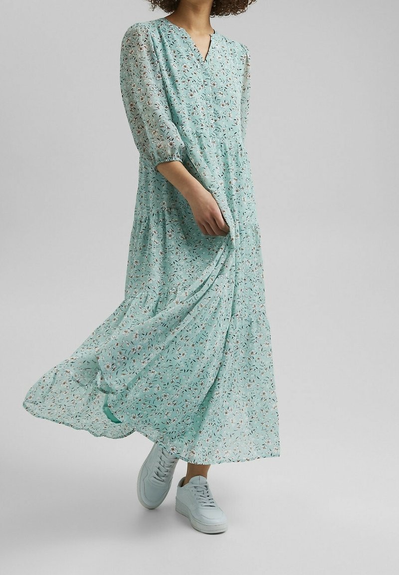 Femme portant une robe fluide bleu clair à motifs floraux et des baskets blanches, tenant la robe en marchant devant un fond uni.