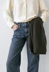 Jasnożółta koszula z guzikami, ciemnoniebieskie jeansy z wysokim stanem i przednimi kieszeniami, do tego ciemnobrązowy sweter zarzucony na ramię.