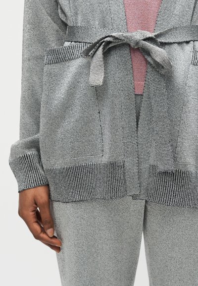 Χέρι που δένει τη ζώνη σε γκρι πλεκτή ζακέτα με εμπρόσθιες τσέπες, φορεμένη πάνω από ροζ τοπ και ταιριαστά γκρι πλεκτά παντελόνια.