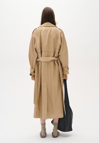 Beige trench coat with a belt, visible buttons, and large lapels. Model holds a black tote bag. Soft fabric, mid-length design.