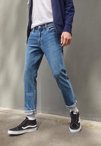 Blå denimjeans med en avsmalnande passform, upprullade ärmar och en femfickdesign, parat med svarta högklackade sneakers och vita strumpor.