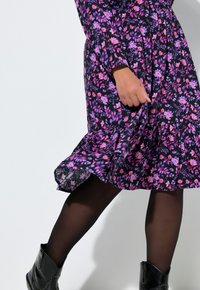 Sukienka w kwiaty w kolorze czarnym z fioletowymi i różowymi wzorami, z długimi rękawami i falbankami na dole, zestawiona z czarnymi botkami za kostkę i kryjącymi rajstopami.