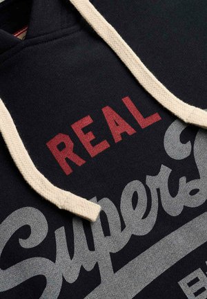 Czarna bluza z kapturem z białymi sznureczkami i czerwono-szarym napisem "REAL Superdry" nadrukowanym na tkaninie.